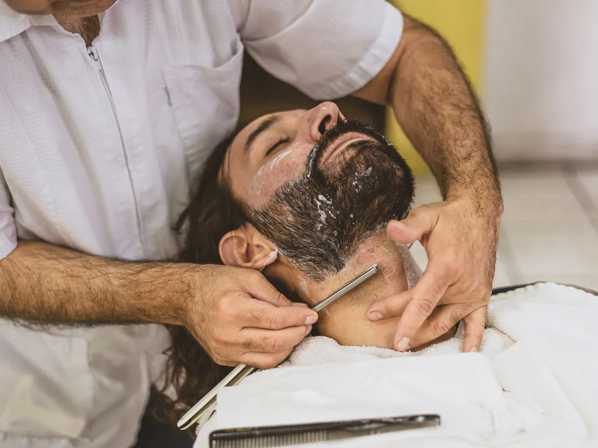 Barbeiro fazendo a barba de um cliente com navalha, com toalha branca no peito